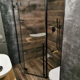 Nowoczesna kabina prysznicowa z czarnymi profilami, drewnopodobne płytki na ścianach i podłodze, widoczna umywalka nablatowa i toaleta.
