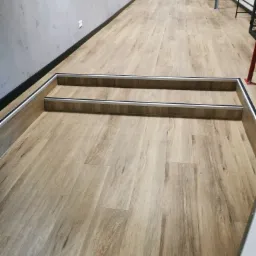 Jasna, drewnopodobna podłoga z panelami, z widocznymi dwoma stopniami schodów zabezpieczonymi aluminiowymi profilami, prowadzącymi w głąb korytarza z szarą ścianą i czerwonymi drzwiami w tle.