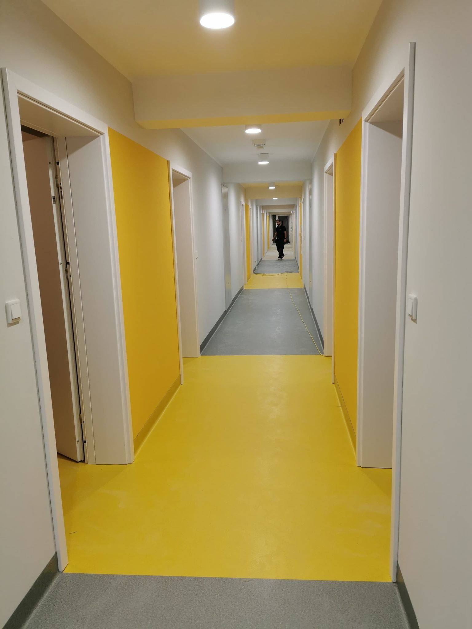 Długi korytarz z jaskrawożółtą podłogą i szarą strefą, białe ściany z żółtymi panelami, białe drzwi, osoba w oddali.