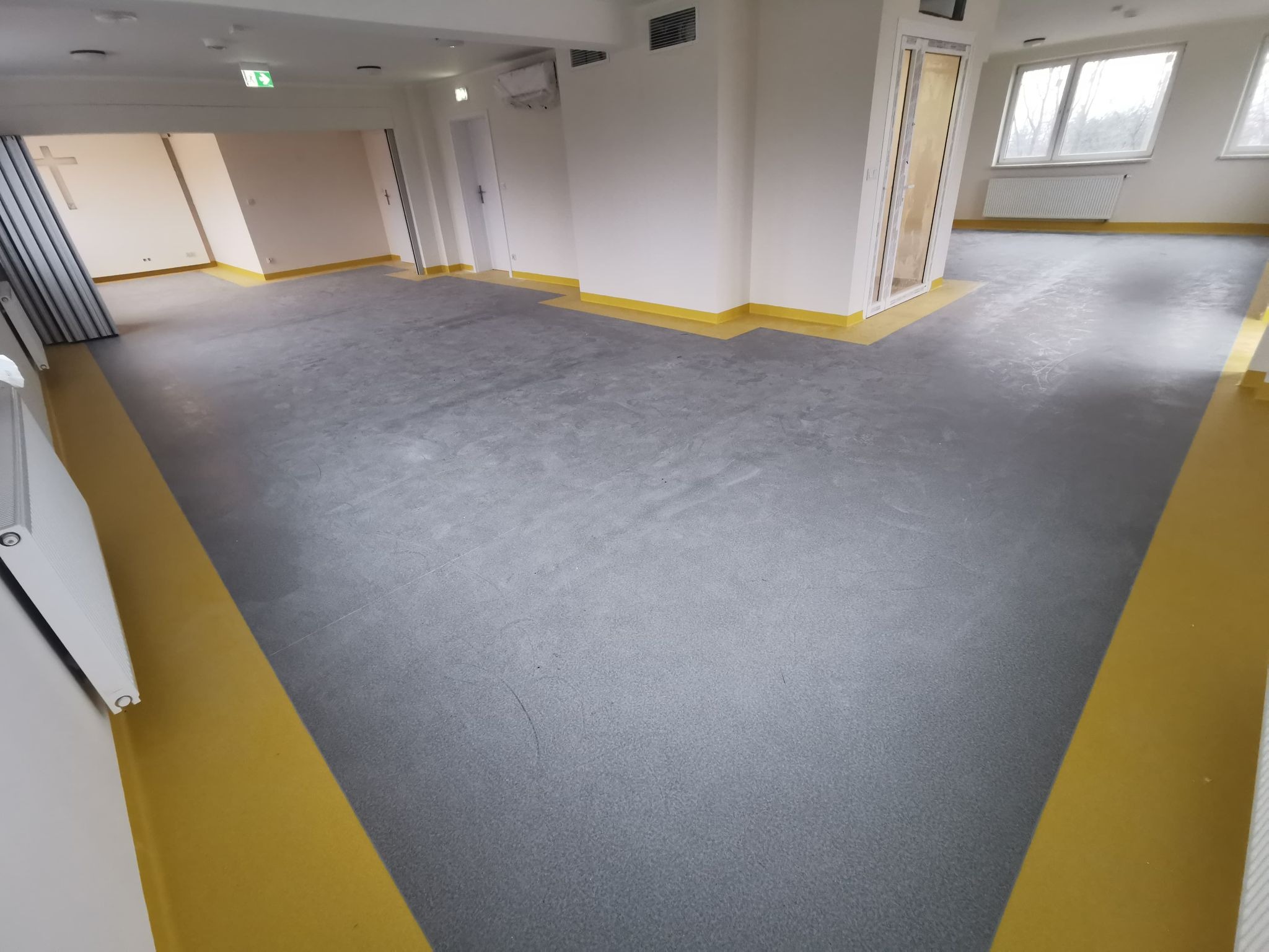 Szara, jednolita podłoga w przestronnym, jasnym pomieszczeniu z żółtymi listwami przypodłogowymi, widok na okna i drzwi w tle.