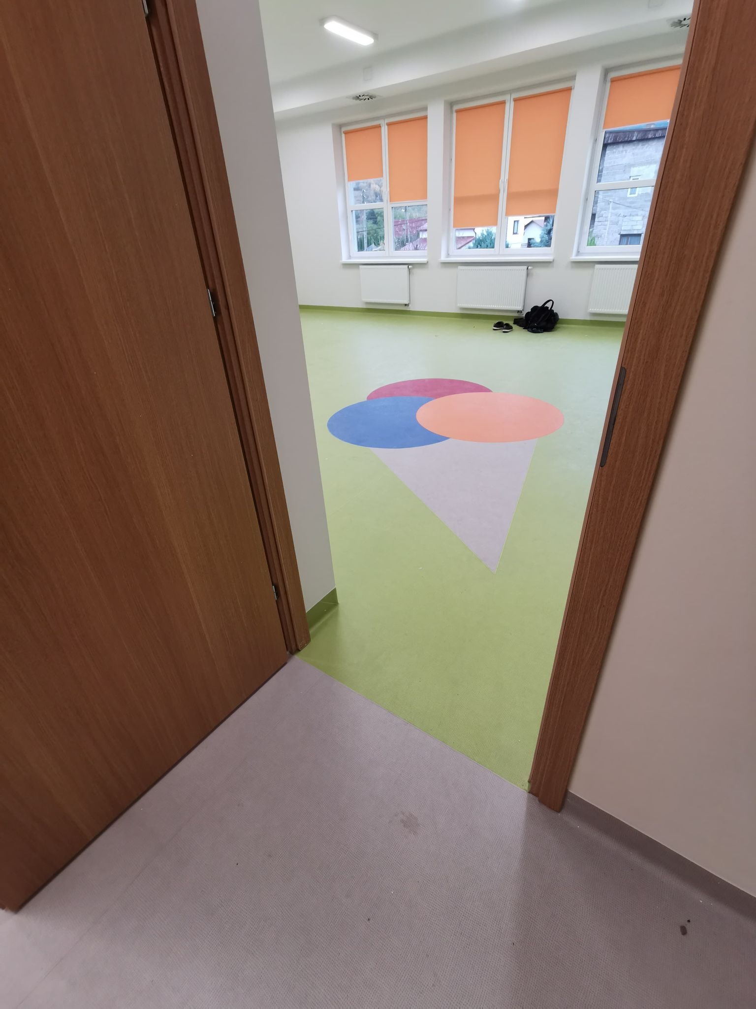 Widok zza uchylonych drzwi na salę z zieloną podłogą, na której widnieje duży, kolorowy wzór lodów w rożku. W tle trzy okna z pomarańczowymi roletami i radiatorami pod nimi. Torba i buty leżą...