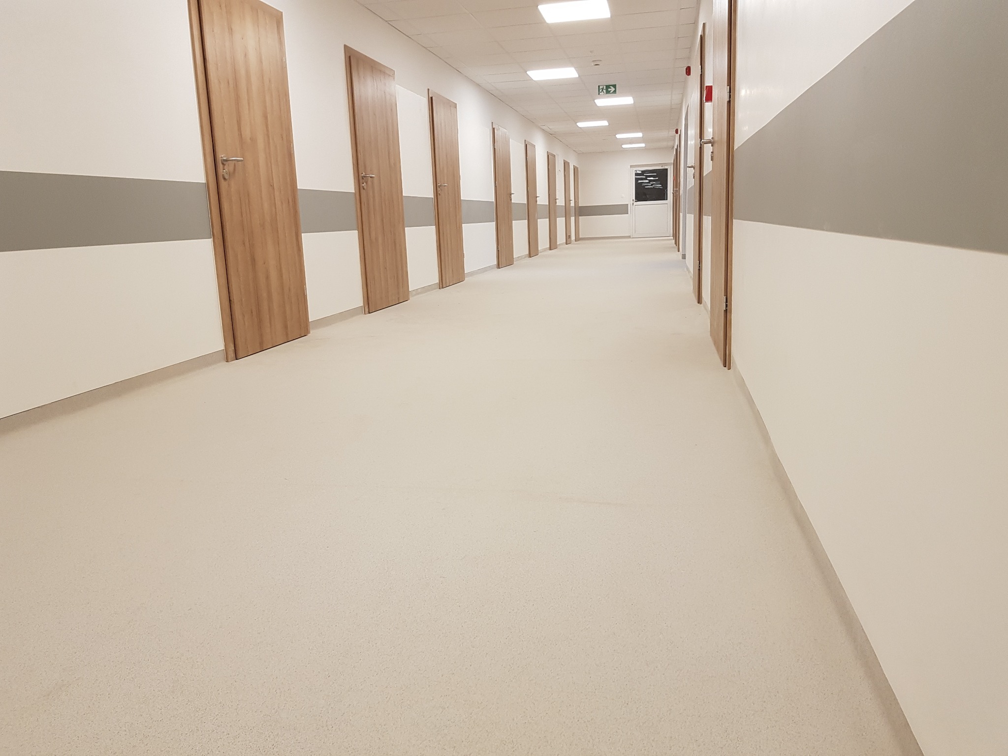 Długi korytarz w budynku użyteczności publicznej z jasną, jednolitą podłogą, drewnopodobnymi drzwiami i szarym pasem na ścianie.