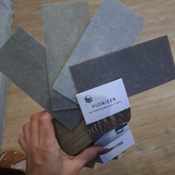 Paweł Mikosz - Próbki heterogenicznego winylu Pioneer w odcieniach szarości i brązu, trzymane w dłoni na tle podłogi, z widoczną kartą producenta Lentex.