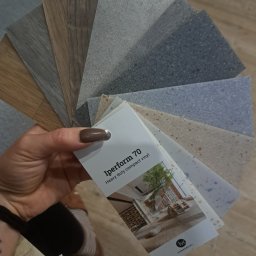 Paweł Mikosz - Prezentacja różnorodnych próbek wykładzin podłogowych w odcieniach szarości i brązu, trzymanych w dłoni z lakierowanymi na brązowo paznokciami, wraz z katalogiem 'Iperform 70 Heavy duty compact...