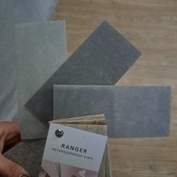 Paweł Mikosz - Próbki heterogenicznego winylu Ranger w odcieniach szarości, ułożone na podłodze imitującej drewno, trzymane w dłoni z lakierowanymi paznokciami.