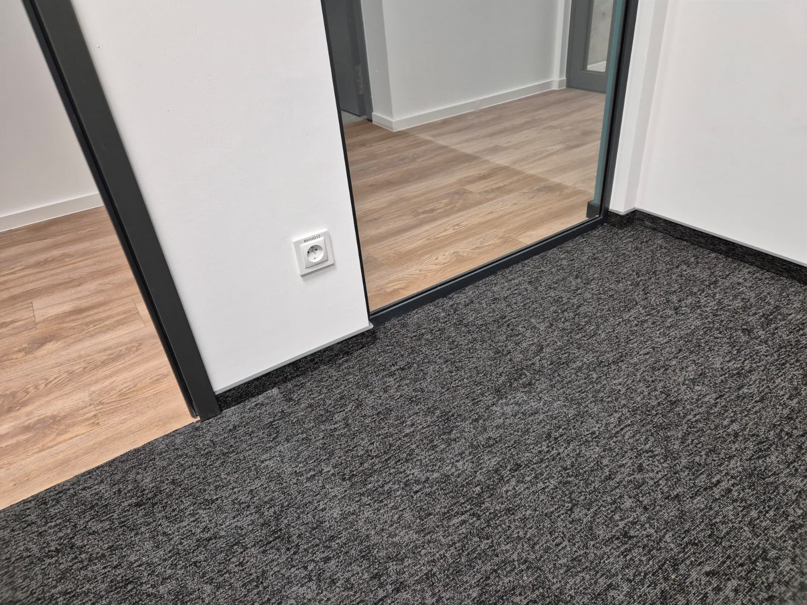 Szara wykładzina dywanowa w biurze, widoczna przy ścianie z gniazdkiem elektrycznym oraz krawędź panelu podłogowego za szklaną ścianką działową.