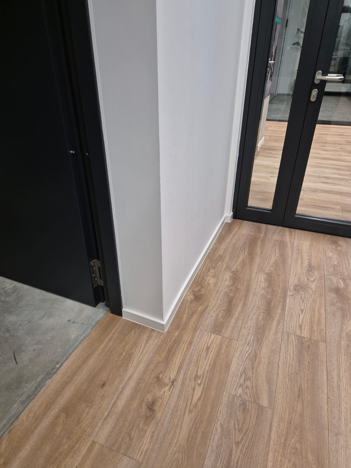Fragment wnętrza biura z panelami podłogowymi o wzorze drewna, białą listwą przypodłogową i czarnymi drzwiami.