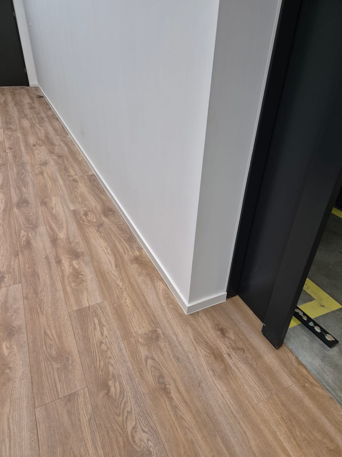 Jasnobrązowe panele podłogowe imitujące drewno, ułożone wzdłuż białej ściany, z widocznym fragmentem czarnej ościeżnicy drzwiowej i szarym fragmentem podłogi technicznej z żółtym oznaczeniem w tle.