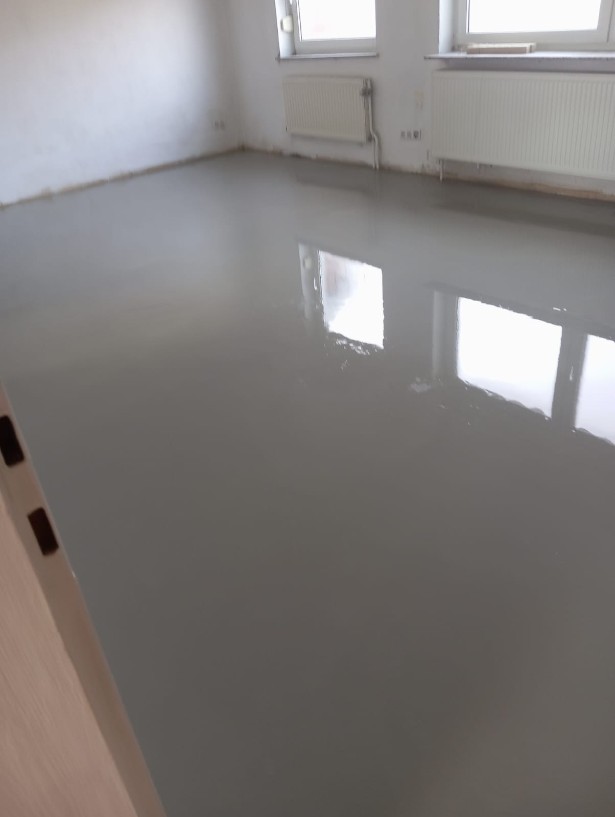Wylana, samopoziomująca szara posadzka w pomieszczeniu z dwoma oknami i grzejnikami, po remoncie w Niedźwiedzy, jeszcze mokra i odbijająca światło.