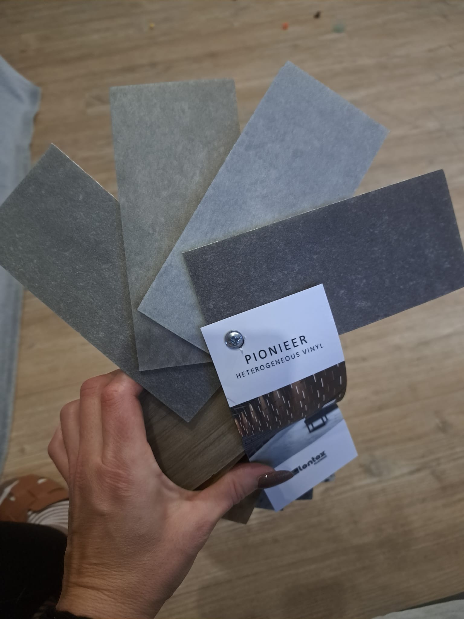 Próbki heterogenicznego winylu Pioneer w odcieniach szarości i brązu, trzymane w dłoni na tle podłogi, z widoczną kartą producenta Lentex.