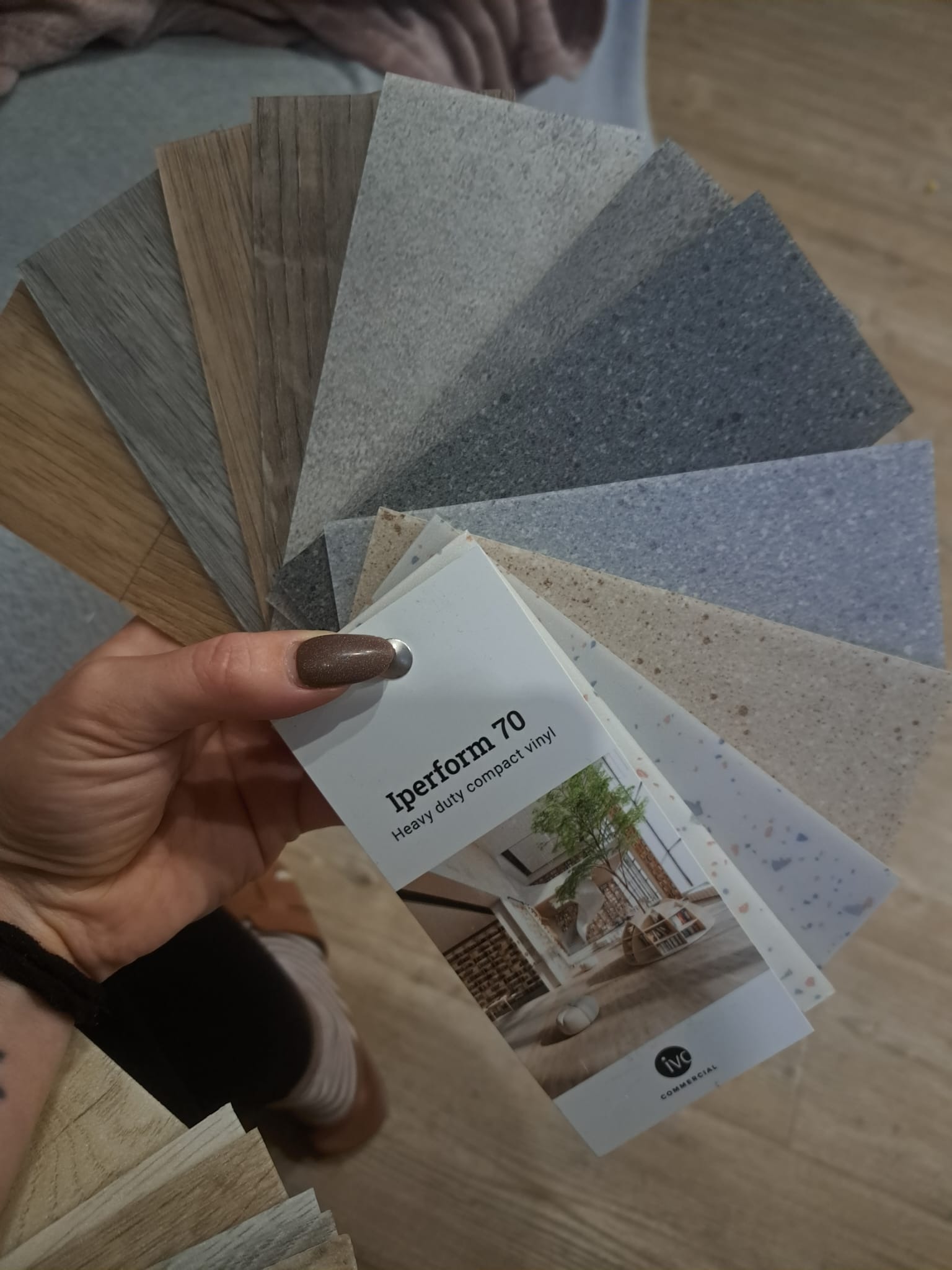 Prezentacja różnorodnych próbek wykładzin podłogowych w odcieniach szarości i brązu, trzymanych w dłoni z lakierowanymi na brązowo paznokciami, wraz z katalogiem 'Iperform 70 Heavy duty compact...