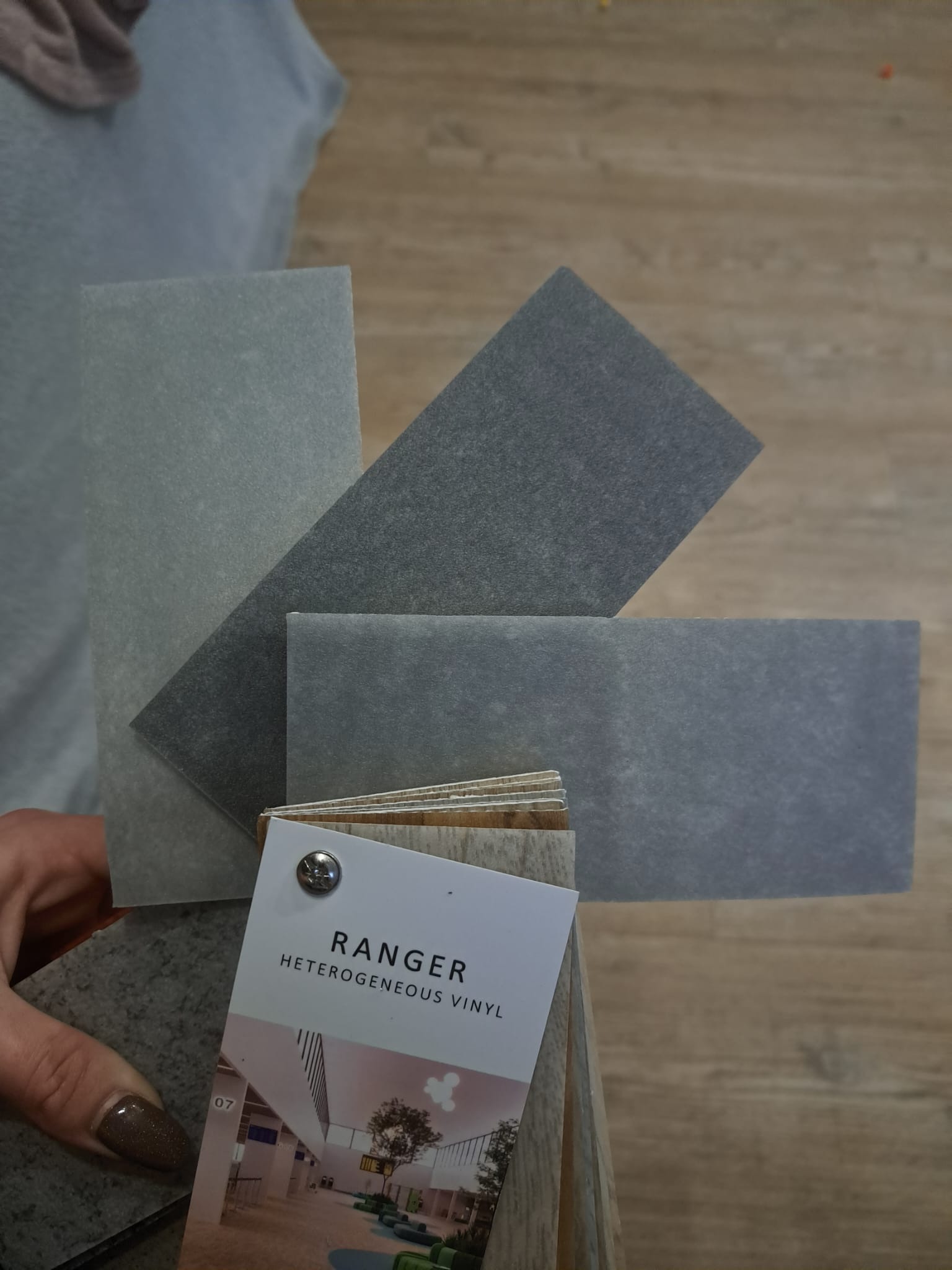 Próbki heterogenicznego winylu Ranger w odcieniach szarości, ułożone na podłodze imitującej drewno, trzymane w dłoni z lakierowanymi paznokciami.