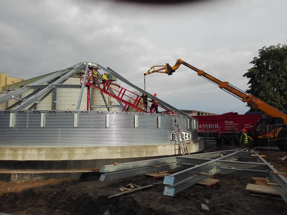 Montaż stalowego silosu zbożowego z użyciem dźwigu, widoczne elementy konstrukcyjne, pracownicy w kaskach i odblaskowych kamizelkach, oraz ciężarówka z napisem ANSROL.