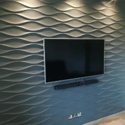 Ściana z panelami 3D w kształcie fal, pomalowana na szaro-niebieski kolor, z zawieszonym telewizorem i soundbarem.