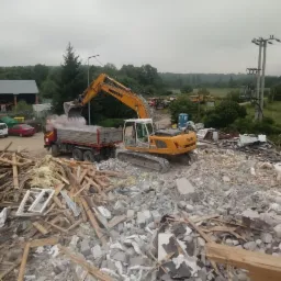 Żółta koparka Liebherr załadowuje gruz do ciężarówki na tle rozbiórki budynku, widoczne fragmenty drewna i betonu.