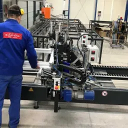 Pracownik w uniformie firmy SAPOR obsługuje automatyczną linię produkcyjną do obróbki profili aluminiowych, prawdopodobnie do produkcji okien i drzwi.