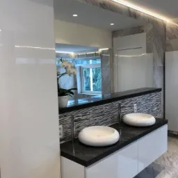 Nowoczesna łazienka z dwoma okrągłymi umywalkami na czarnym blacie, białe szafki pod umywalkami, lustro z oświetleniem LED, marmurowe płytki na ścianach, kwiat w doniczce odbity w lustrze.