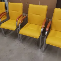 Rząd czterech żółtych krzeseł biurowych z drewnianymi podłokietnikami i chromowanymi nogami, ustawionych w warsztacie lub magazynie.