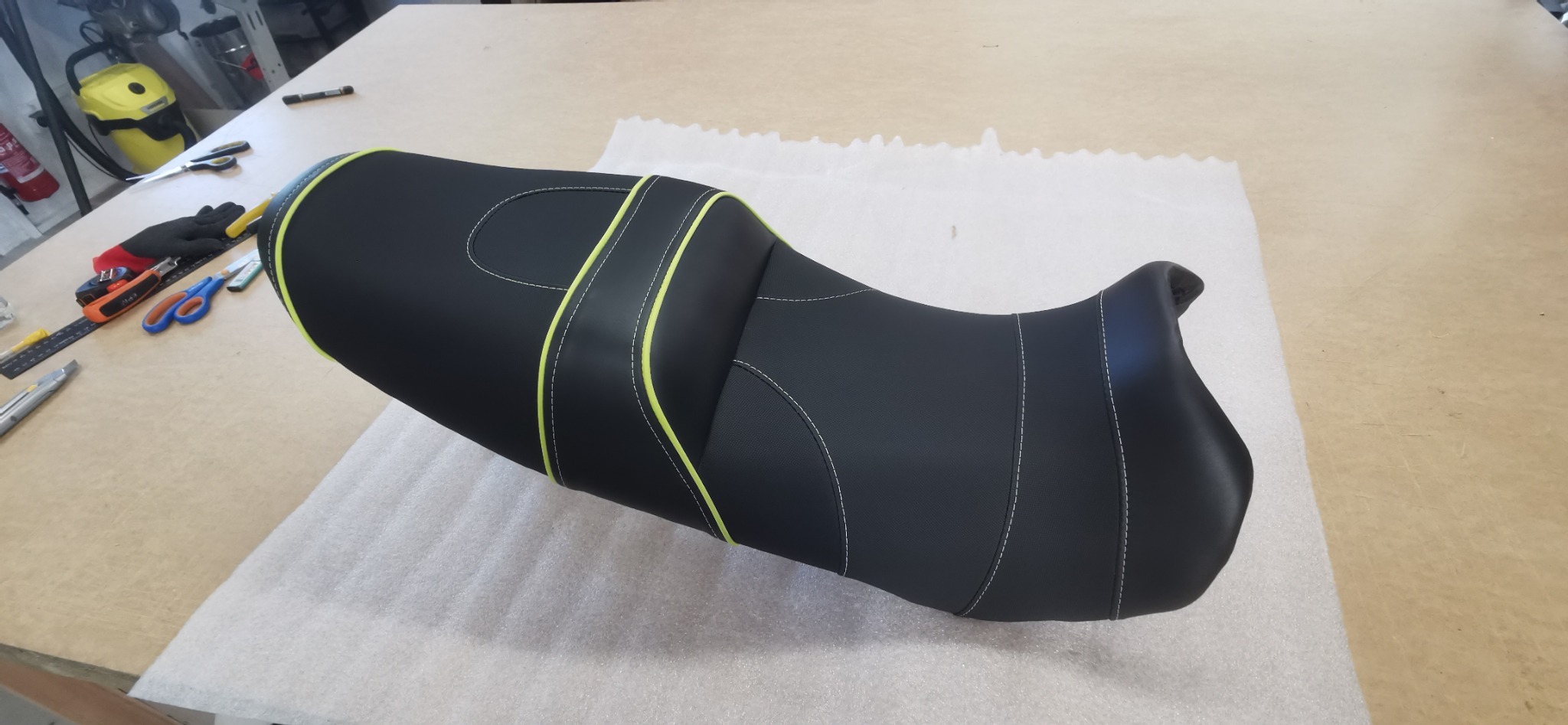 Odnowione czarne siedzisko motocyklowe z jaskrawożółtymi akcentami i białymi szwami, leżące na stole warsztatowym obok narzędzi.