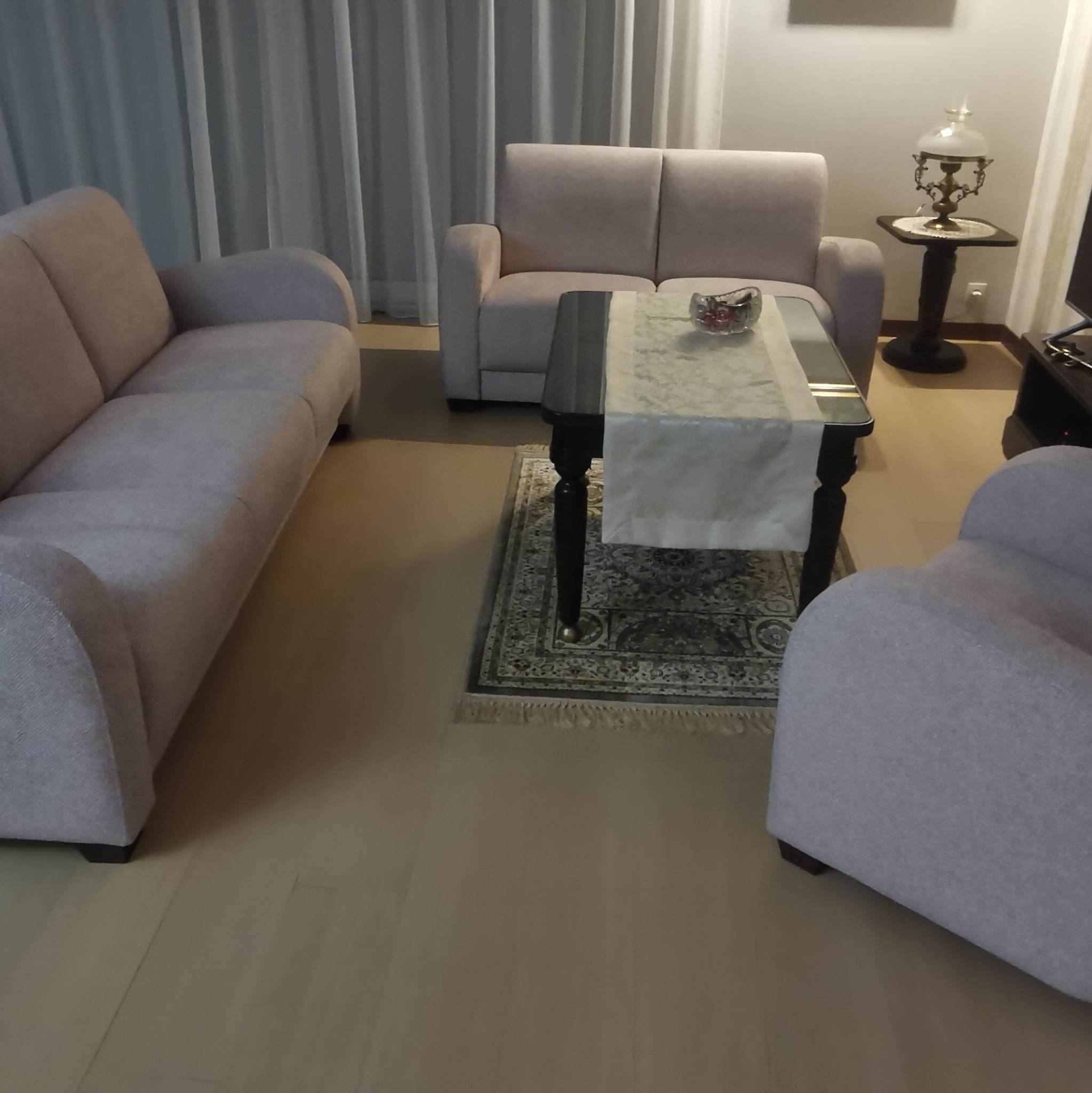 Jasne, tapicerowane meble wypoczynkowe w salonie: trzyosobowa sofa, dwuosobowa sofa i fotel, ustawione wokół czarnego stolika kawowego z bieżnikiem i dekoracyjną misą, na tle zasłon.