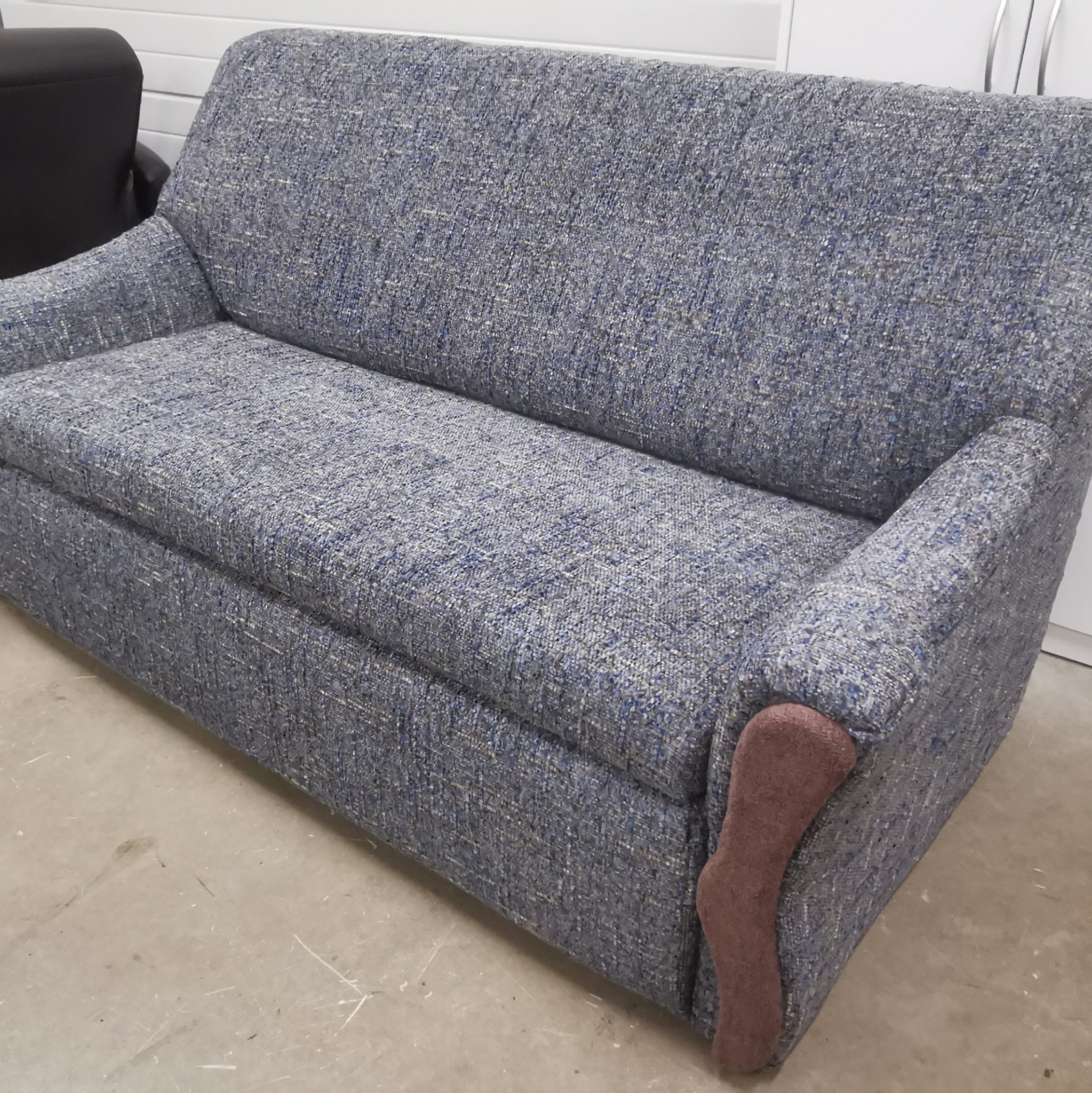 Dwuosobowa sofa z szaro-niebieską tapicerką o wyraźnej fakturze, z brązowym elementem dekoracyjnym na jednym z podłokietników, stojąca na betonowej podłodze.