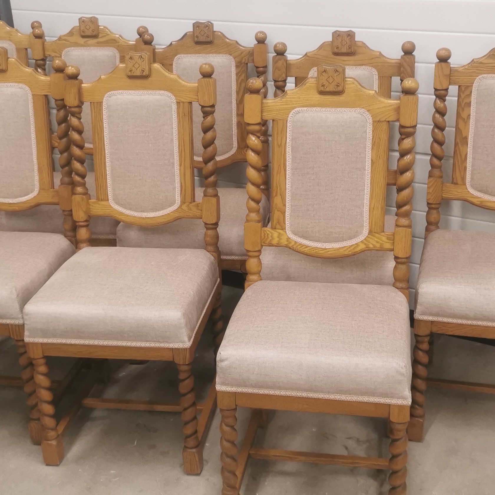Rząd odnowionych krzeseł dębowych z tapicerowanymi siedziskami i oparciami w neutralnym beżowym odcieniu, z charakterystycznymi toczonymi nogami i zdobieniami na szczycie oparcia.