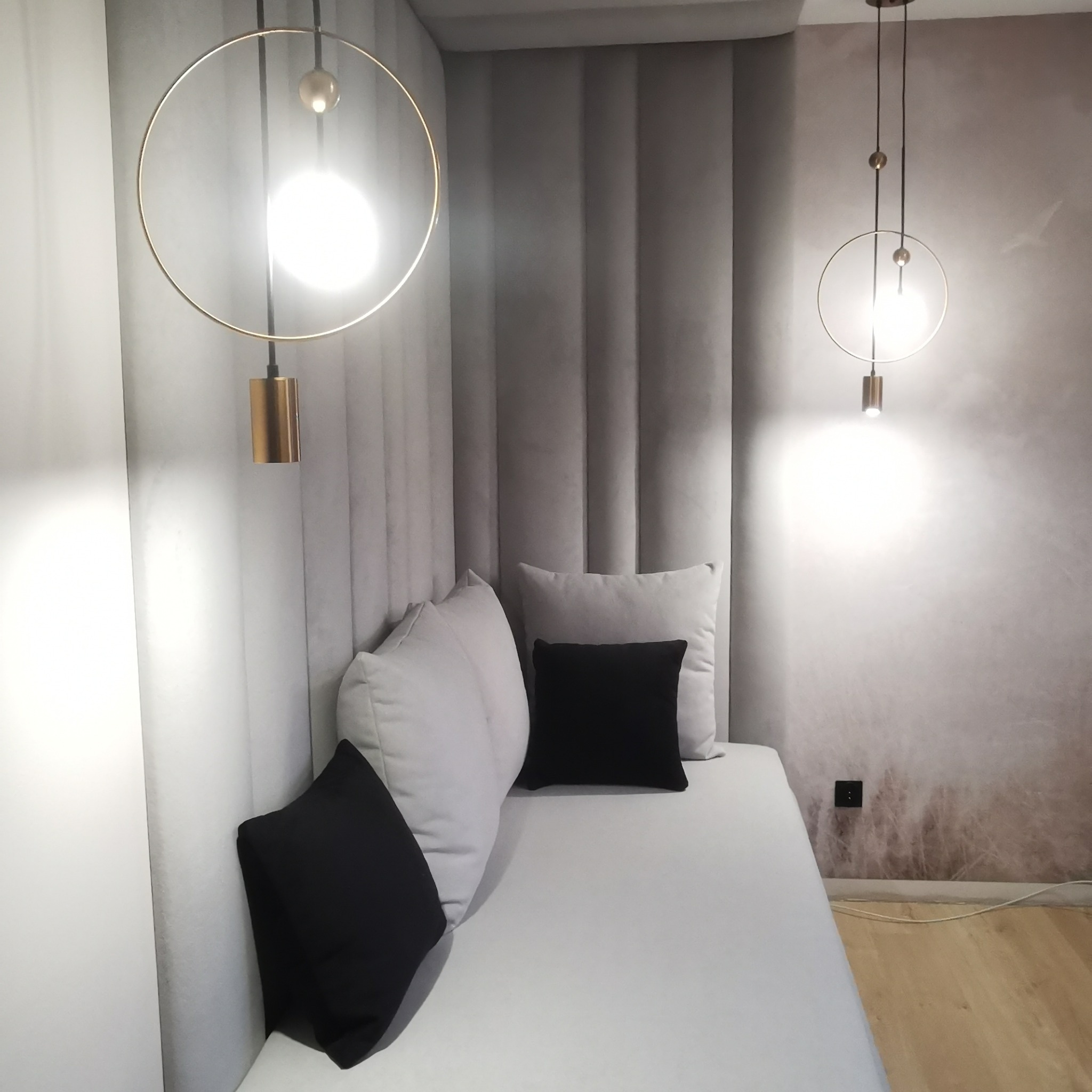 Wykończona wnęka z siedziskiem i tapicerowanym panelem ściennym w odcieniach szarości, oświetlona dwoma nowoczesnymi lampami wiszącymi z okrągłymi kloszami i złotymi elementami.