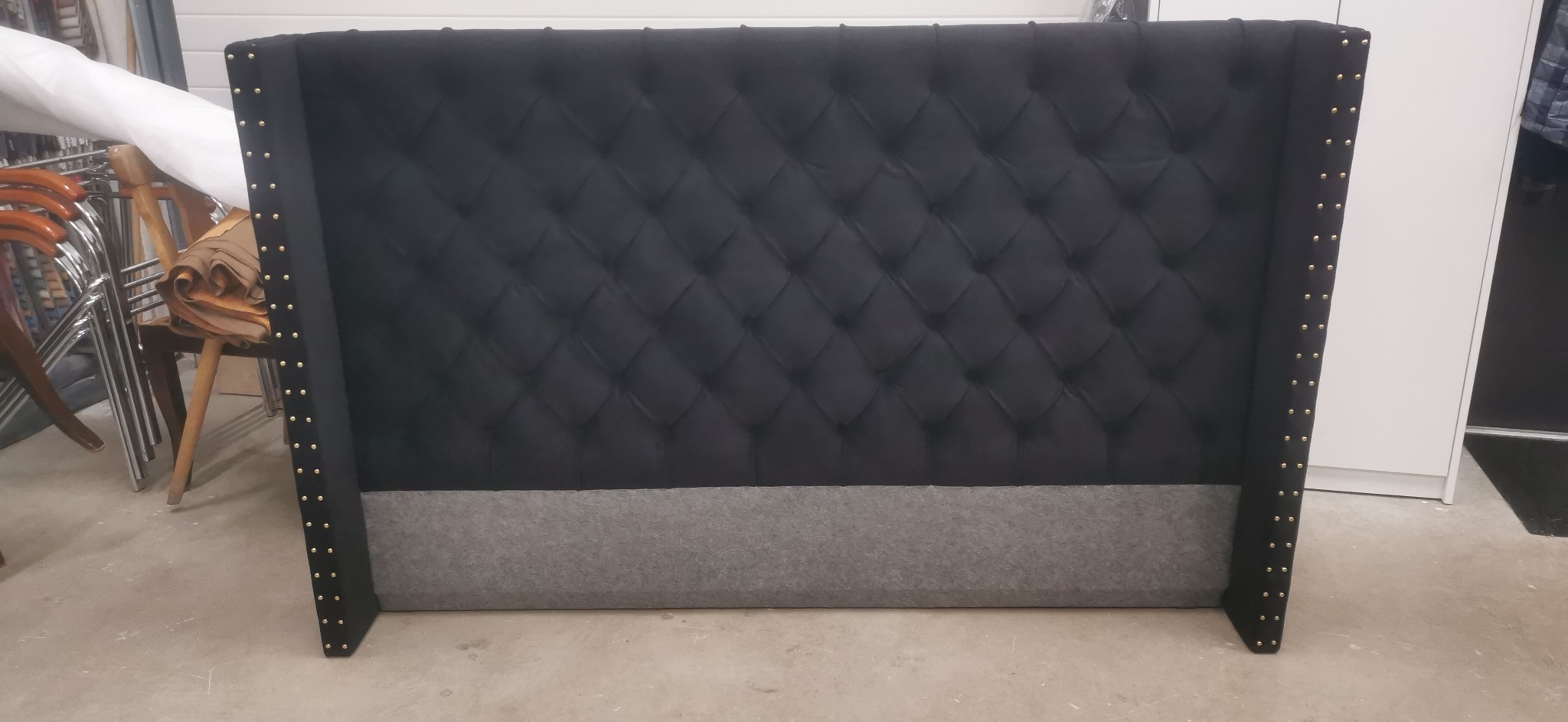 Czarne, pikowane wezgłowie łóżka z szarym pasem u dołu i ozdobnymi, złotymi nitami po bokach, sfotografowane w warsztacie.