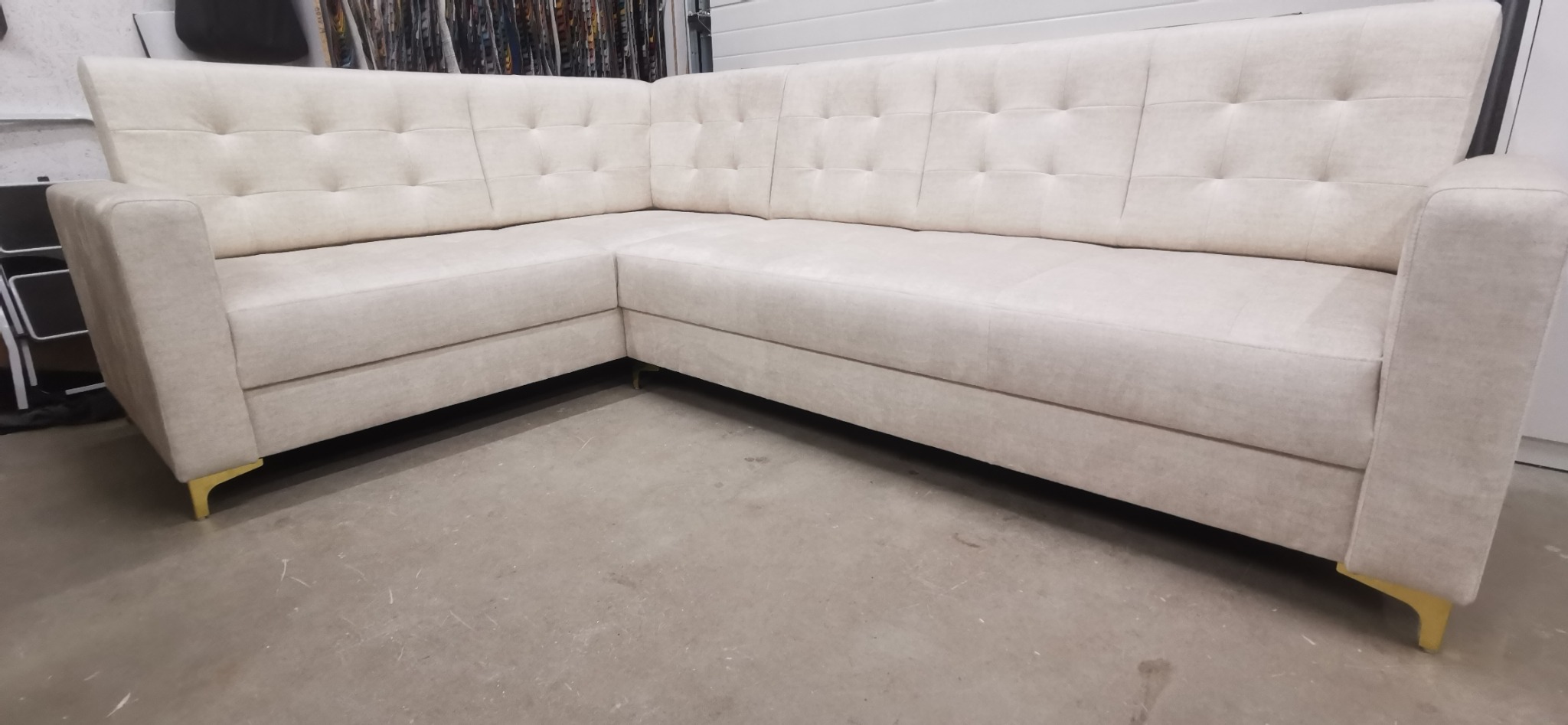 Jasnoszary narożnik z pikowanym oparciem i złotymi nóżkami, widok z przodu na szarym tle podłogi.