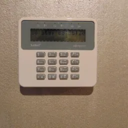 Biały panel sterowania systemem alarmowym marki Satel Perfecta zamontowany na beżowej ścianie, widoczny wyświetlacz LCD z datą i godziną oraz klawiatura numeryczna z przyciskami funkcyjnymi.