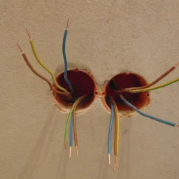 Dwa otwory w ścianie z wystającymi przewodami elektrycznymi w kolorach niebieskim, żółto-zielonym i brązowym, przygotowane do podłączenia gniazdka lub włącznika.