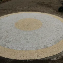 Okrągły plac wyłożony kostką brukową w dwóch odcieniach, otoczony przygotowaną ziemią pod przyszły trawnik i rośliny.