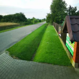 Widok z góry na świeżo założony trawnik z systemem nawadniania wzdłuż drogi, z widoczną kostką brukową i ogrodzeniem z reklamą firmy.