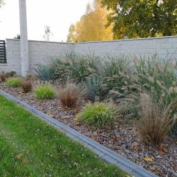 Ogrody Patryk Obrębski - Kaskadowe trawy ozdobne i juki posadzone wzdłuż muru z betonowych bloczków, kora rozsypana jako ściółka, obrzeże z szarej kostki betonowej, trawnik w tle.