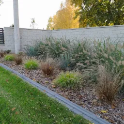 Kaskadowe trawy ozdobne i juki posadzone wzdłuż muru z betonowych bloczków, kora rozsypana jako ściółka, obrzeże z szarej kostki betonowej, trawnik w tle.