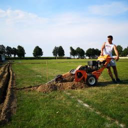 Ogrody Patryk Obrębski - W Pułtusku, ogrodnik obsługuje pomarańczową glebogryzarkę, wykopującą wąski rów na trawiastym boisku, przygotowując teren pod system nawadniania.