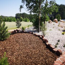 Ogrody Patryk Obrębski - Nowo założony ogród z korą, kamieniami i niską roślinnością, wykończony czerwoną cegłą, z widokiem na pole i las w tle.