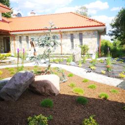 Ogrody Patryk Obrębski - Nowo założony ogród z młodymi roślinami, korą, kamieniami i systemem nawadniania na tle domu z czerwoną dachówką; widoczna osoba w tle