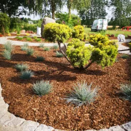 Starannie zaprojektowany ogród z ozdobnymi krzewami, trawami i korą, obrzeżony kamienną ścieżką, z widocznym w tle samochodem dostawczym firmy ogrodniczej.