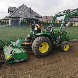 Ogrody Patryk Obrębski - Zielony traktor John Deere z żółtymi felgami, wyposażony w glebogryzarkę i ładowarkę, przygotowuje ziemię pod trawnik na tle ogrodzenia z siatki i domów.