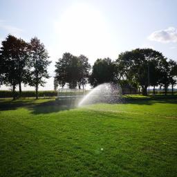 Ogrody Patryk Obrębski - Automatyczny zraszacz nawadnia zielony trawnik na tle drzew i małej budowli, tworząc tęczę w promieniach słońca.