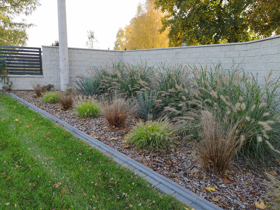 Kaskadowe trawy ozdobne i juki posadzone wzdłuż muru z betonowych bloczków, kora rozsypana jako ściółka, obrzeże z szarej kostki betonowej, trawnik w tle.