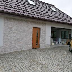 NOWY DOM Anna Serzysko - Wykończony dom z elewacją z kamienia dekoracyjnego, brązowymi drzwiami wejściowymi i żółtym vanem firmy budowlanej na brukowanym podjeździe.