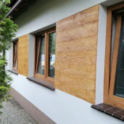 Elewacja domu z oknami, wykończona tynkiem dekoracyjnym imitującym drewno, widok z zewnątrz, z fragmentem iglastego drzewa po lewej stronie.