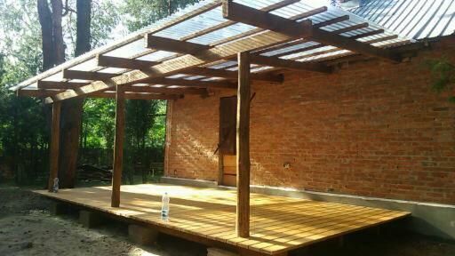 Zadaszony drewniany taras przylegający do murowanej ściany budynku, z przezroczystym zadaszeniem i butelką wody na podłodze.