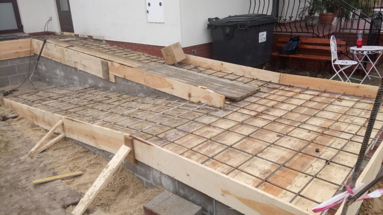 Przygotowanie zbrojenia i szalunku pod wylewkę betonową na zewnątrz budynku, widoczne drewniane deski, siatka zbrojeniowa i piasek.