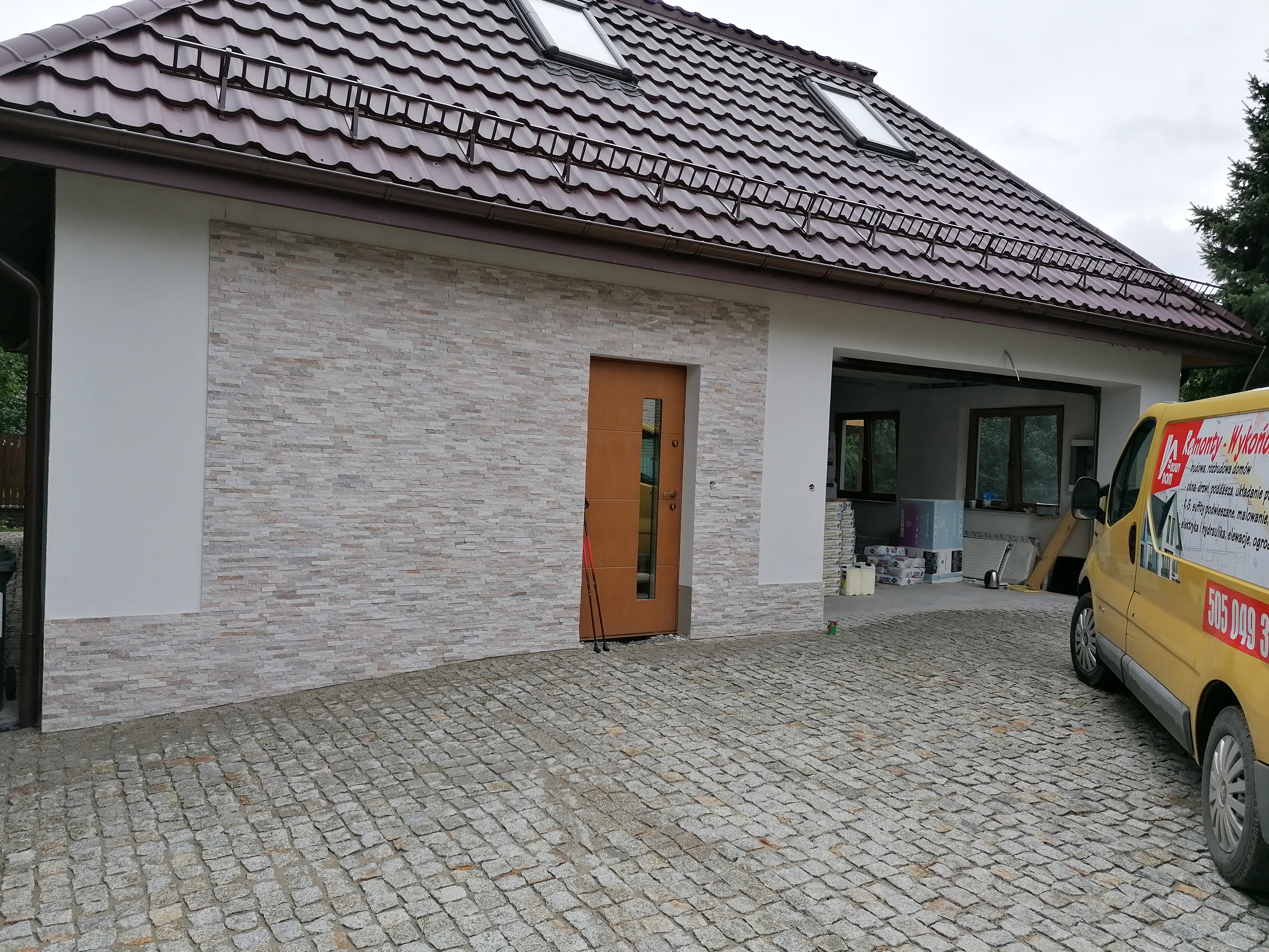 Wykończony dom z elewacją z kamienia dekoracyjnego, brązowymi drzwiami wejściowymi i żółtym vanem firmy budowlanej na brukowanym podjeździe.