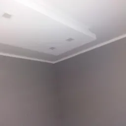 Szare ściany w rogu pokoju z białym, podwieszanym sufitem z wbudowanymi, kwadratowymi lampami LED i białymi listwami przypodłogowymi.