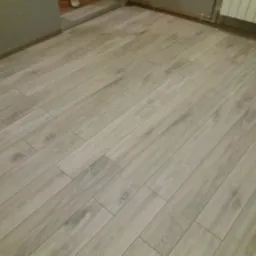 Ułożona pod kątem jasna podłoga z paneli imitujących drewno, widoczne fragmenty ścian i grzejnika.