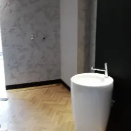 Nowoczesna łazienka z umywalką kolumnową, parkietem w jodełkę i ścianą z efektem dekoracyjnym. Czarny żyrandol nadaje wnętrzu elegancji.